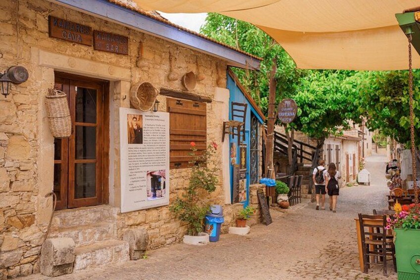 A stone building with a sign reading 'KAMARAS BAR'. There are people walking on the cobblestone street, and a blue awning is visible. 1