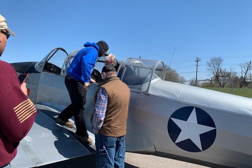 A vintage military aircraft with a star insignia, a person in a blue jacket climbing onto it, and another person standing nearby. 4