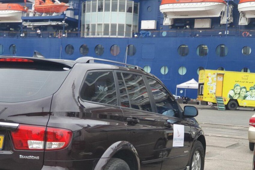 A dark-colored SUV parked in front of a large blue ship with multiple windows and a yellow truck with a mural in the background. 3