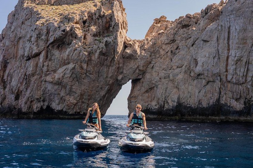 Two people jet-skiing near a large rock formation in the water. 1