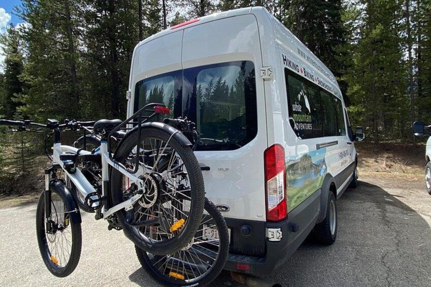 A white van with a bicycle rack attached to the back, parked in a forested area. 3