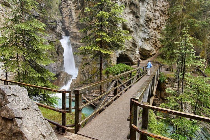 A wooden bridge over a waterfall, surrounded by trees and rocky cliffs. 1