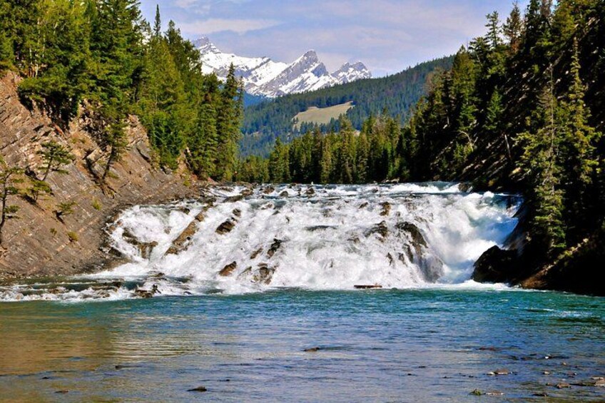 A waterfall cascading into a river surrounded by dense forest and snow-capped mountains in the background. 2