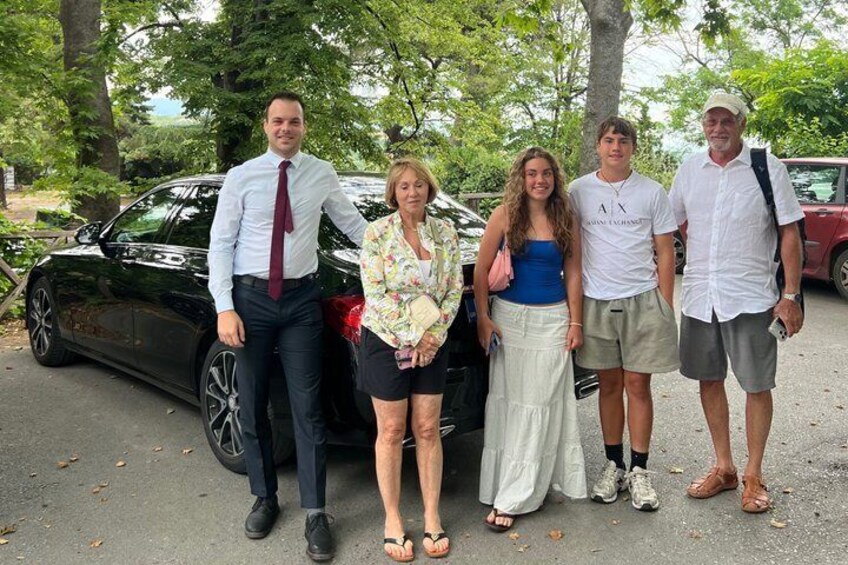 A group of people standing in front of a parked car with trees in the background. 2