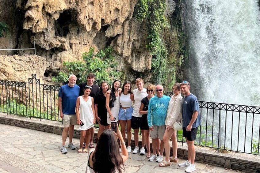 A group of people standing by a waterfall with a rocky cliff in the background. 3