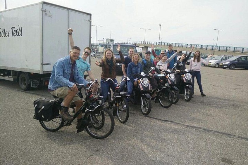 A group of people on bicycles and motorcycles in a parking lot with a truck labeled 'Solex Texel' in the background. 2