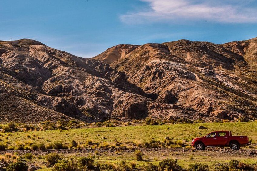 A red pickup truck on a dirt road in a mountainous area with green grass and rocky terrain. 5