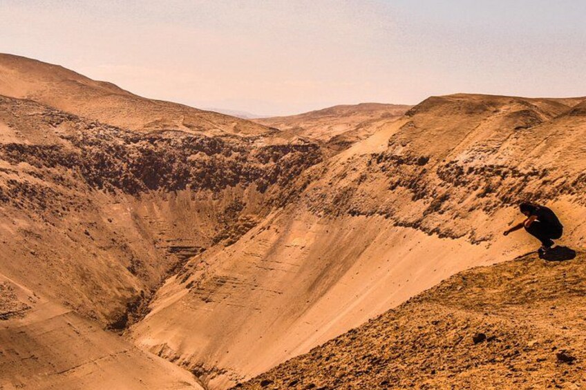 A person crouching on a hill overlooking a valley with sparse vegetation and a clear sky. 4