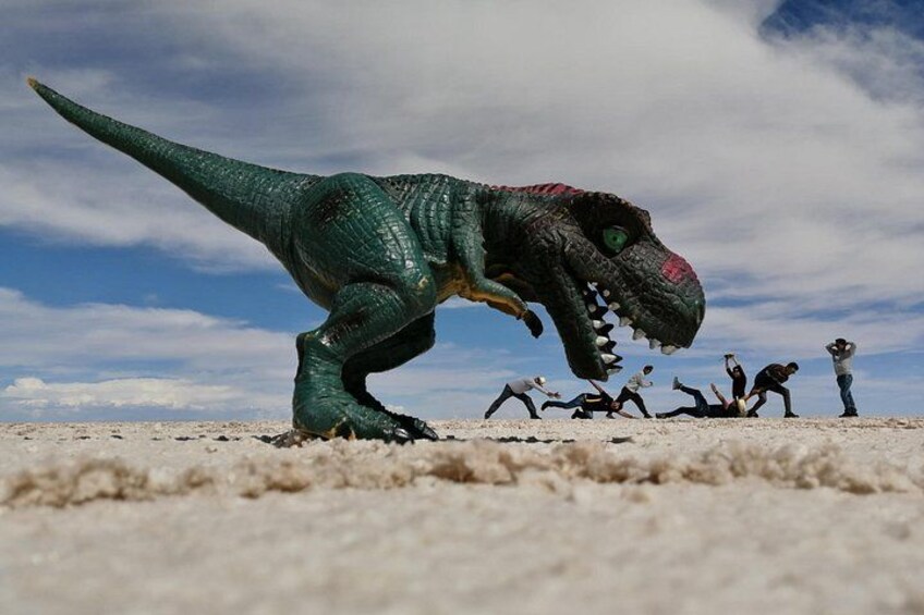 A toy dinosaur on a sandy surface with a group of people in the background. 5