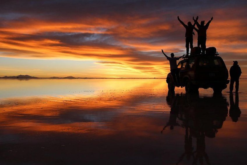 A sunset over a calm body of water with silhouettes of people and a vehicle. 1