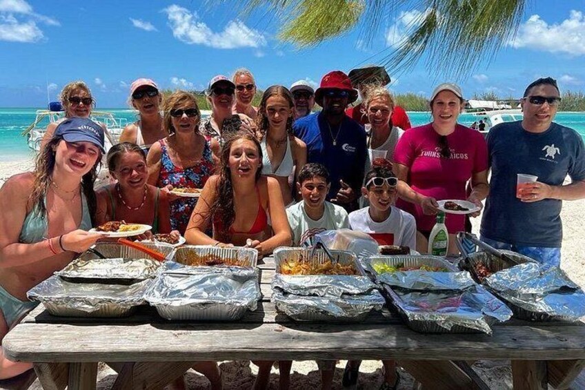 Un grupo de personas reunidas alrededor de una mesa de madera en la playa, con comida y bebidas. 4