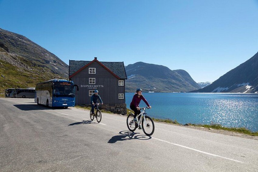 Two cyclists on a road with a bus and a building labeled 'Djupvasshytte' in the background. 2