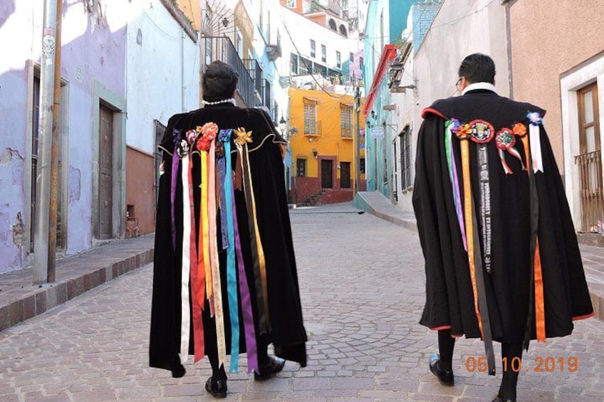 Twee personen lopen over een geplaveid straatje, elk gekleed in een zwarte robe versierd met kleurrijke linten. 5