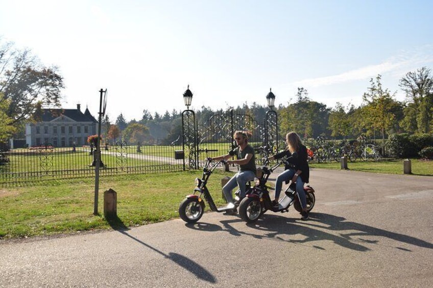 Two people riding electric bikes on a paved path in a park with a fence, lamp posts, and a building in the background. 3