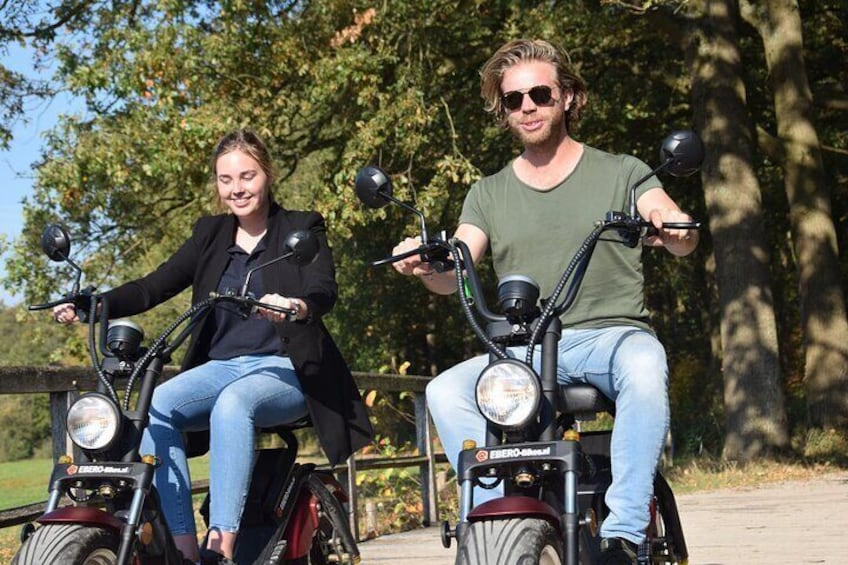 Two people riding electric scooters on a paved path with trees in the background. 4