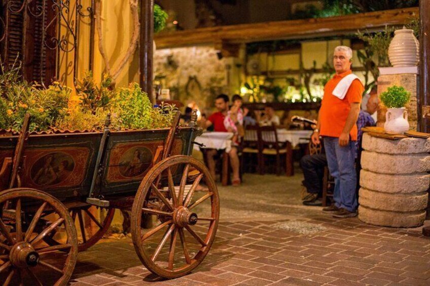Un espacio de comedor al aire libre rústico, con un carrito de madera lleno de plantas, un hombre con una camisa naranja y una jardinera de piedra con planta en maceta. 3