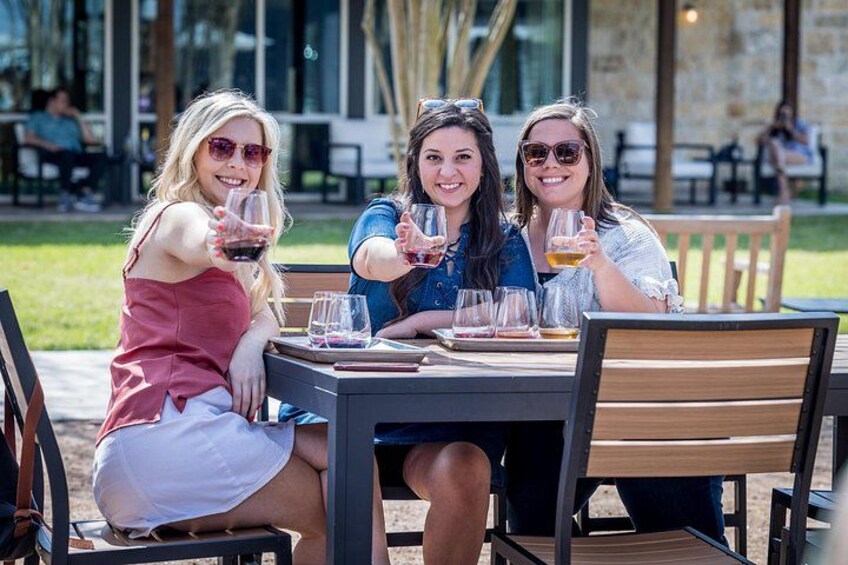 Trois femmes sont assises à une table à l’extérieur, devant des verres à vin, dans un cadre ensoleillé avec des chaises et des tables. 4