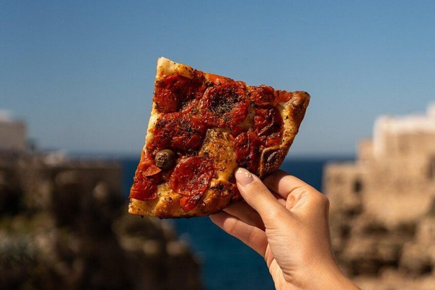 Una porción de pizza que sostiene una mano, con un paisaje costero de fondo. 2