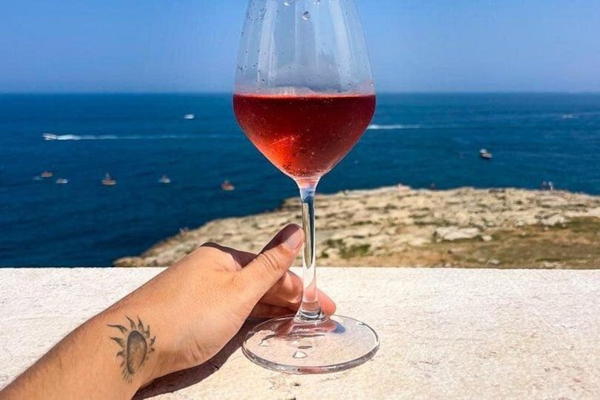 Una mano sosteniendo una copa de vino con líquido rojo, un tatuaje en la muñeca y un paisaje costero con mar y barcos al fondo. 2