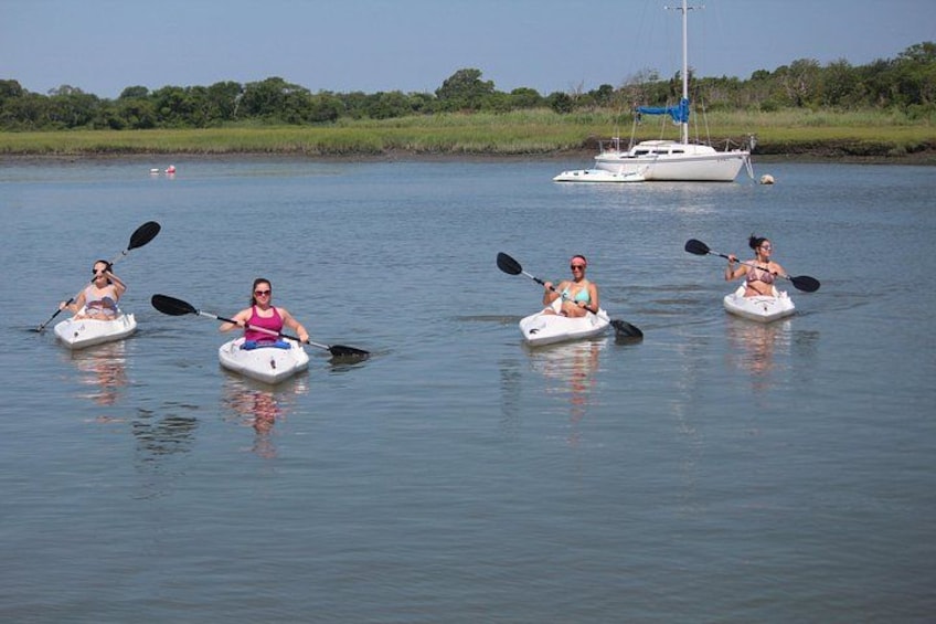 Four people kayaking on a calm river with a sailboat in the background. 1
