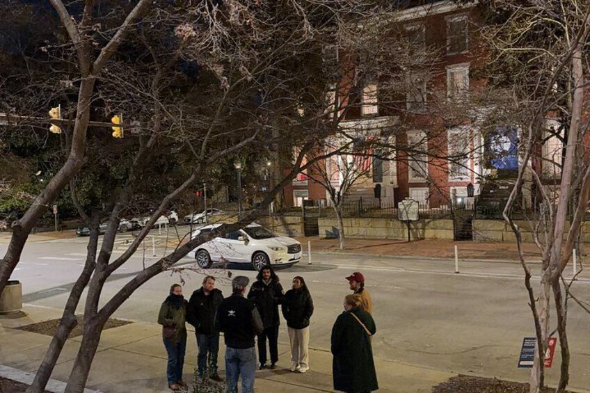 A group of people standing on a sidewalk, a white car parked on the street, and a building with a red brick facade in the background. 4