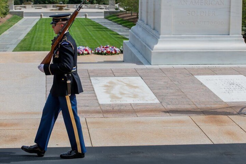 A military personnel in uniform standing at attention in front of a monument with inscriptions. 1
