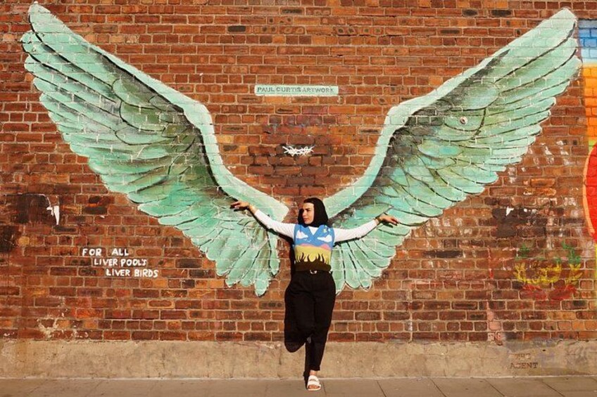 A person standing in front of a mural with large green wings and text reading 'FOR ALL LIVER POOLS LIVER BIRDS'. 1