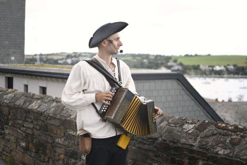 A person playing a traditional accordion outdoors, dressed in period costume, with a stone wall and a body of water in the background. 2