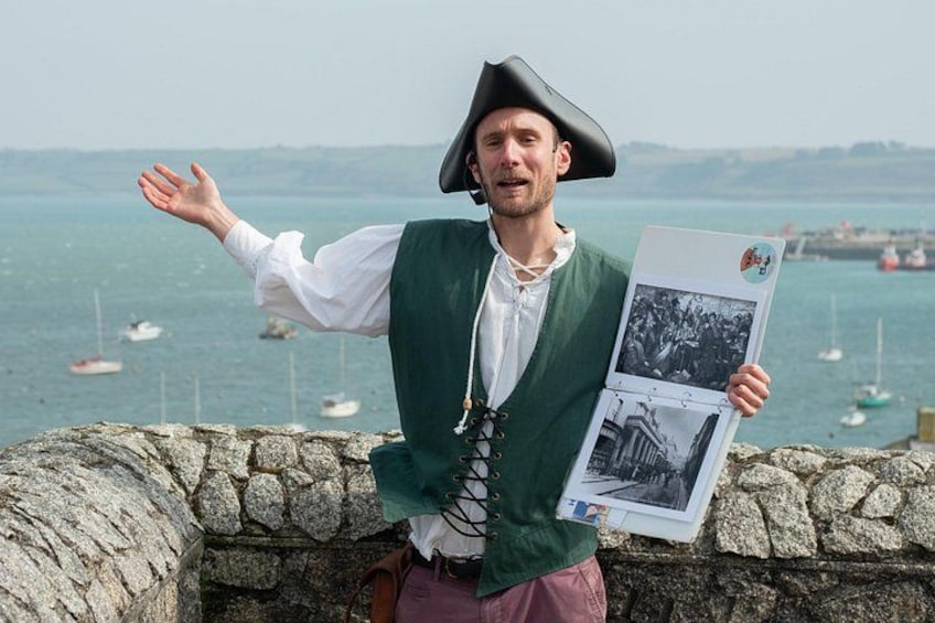 A person in historical costume holding a booklet with a photograph, standing by a stone wall with a view of the sea and boats. 2