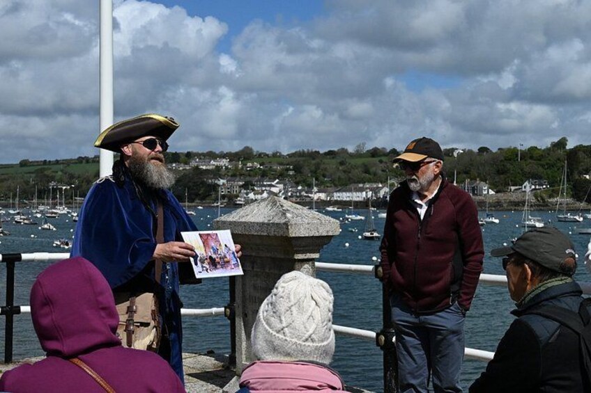 A man in a blue jacket and hat standing by a body of water with boats, a stone structure, and a town in the background. 2