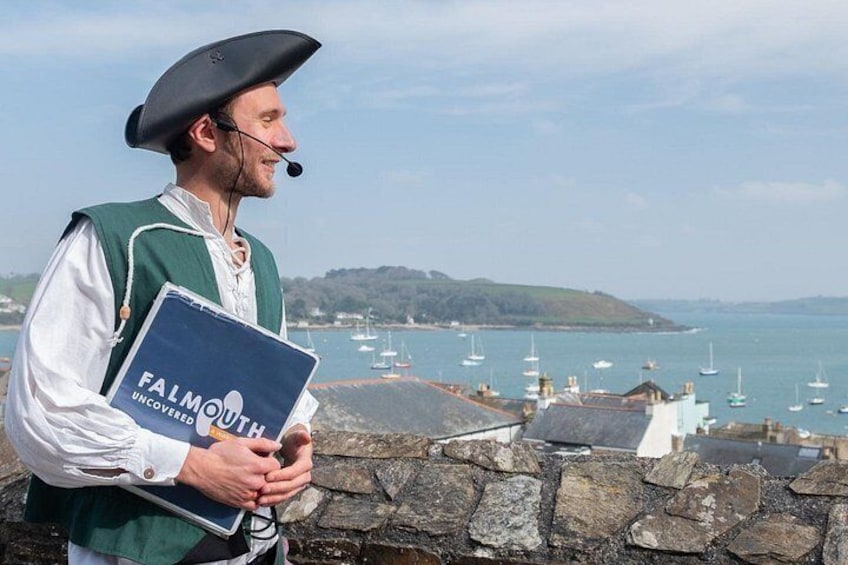 A person in historical attire holding a book titled 'Falmouth Uncovered' by a stone wall with a view of boats and hills in the background. 1