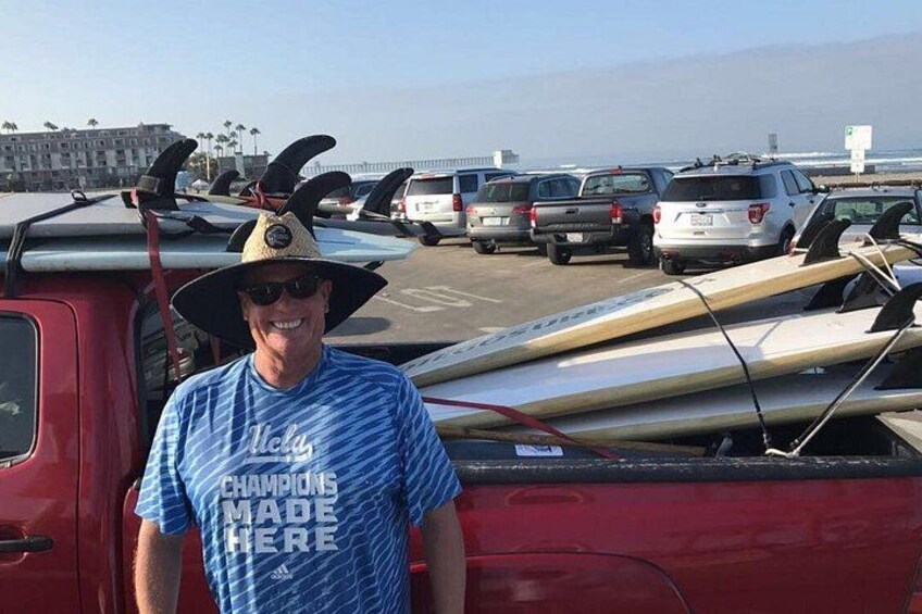 A person standing next to a red truck with surfboards on top, parked in a lot with other vehicles and a beach in the background. 1