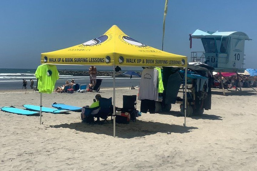 Una spiaggia con una tenda gialla che pubblicizza lezioni di surf, sedie a sdraio e tavole da surf. 2