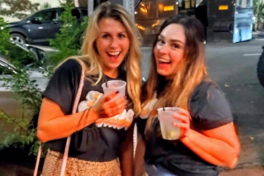 Two people holding drinks, standing on a sidewalk with cars and greenery in the background. 4
