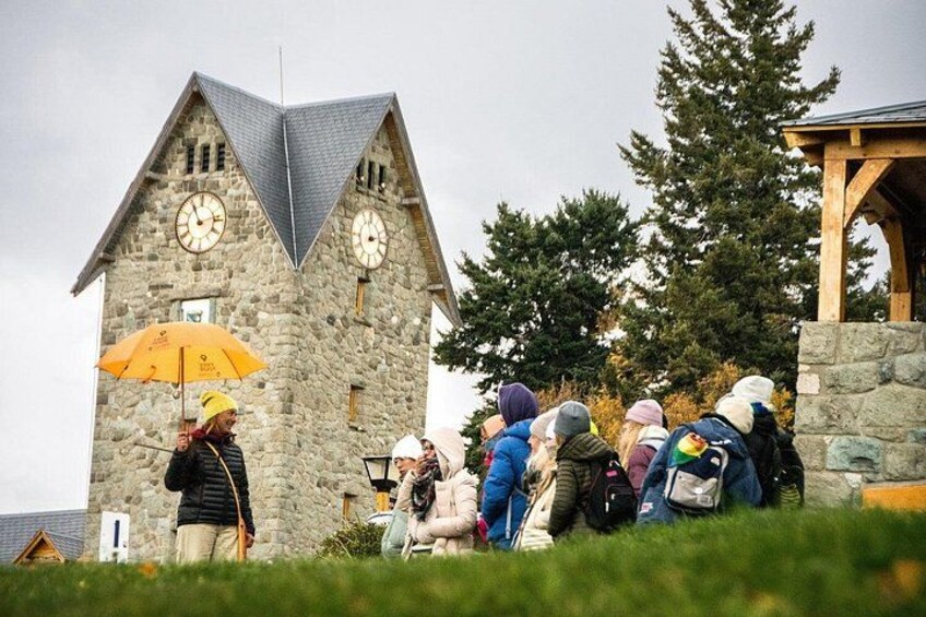 Un edificio in pietra con tetto a forma di triangolo e due orologi, circondato da persone e alberi. 5