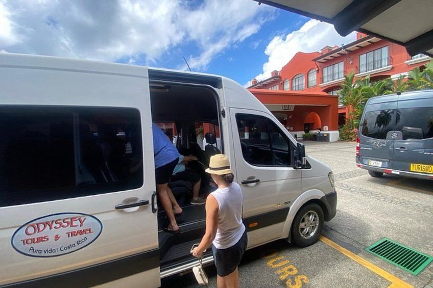 Una camioneta blanca con la leyenda 'ODYSSEY TOURS & TRAVEL' y 'Pura Vida Costa Rica', estacionada en un estacionamiento con un edificio al fondo. 3