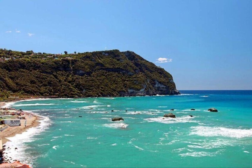 A coastal landscape with turquoise waters, a sandy beach, and a hill with vegetation. 1