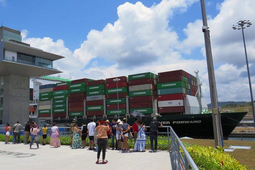 A ship with stacked shipping containers, a crowd of people, and a modern building in the background. 2