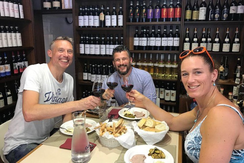 Three people toasting with wine glasses at a table with food and bottles of wine in the background. 2