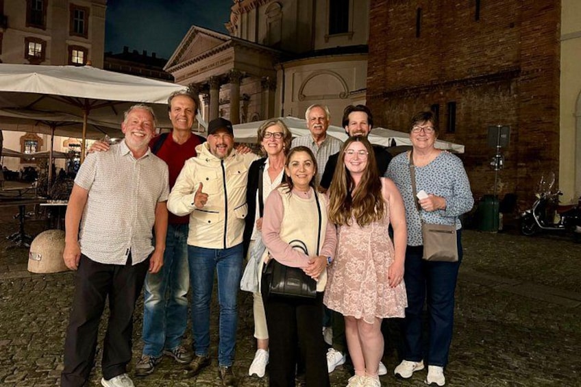 A group of people standing in an outdoor setting with cobblestone pavement, a brick building, and a white umbrella in the background. 4