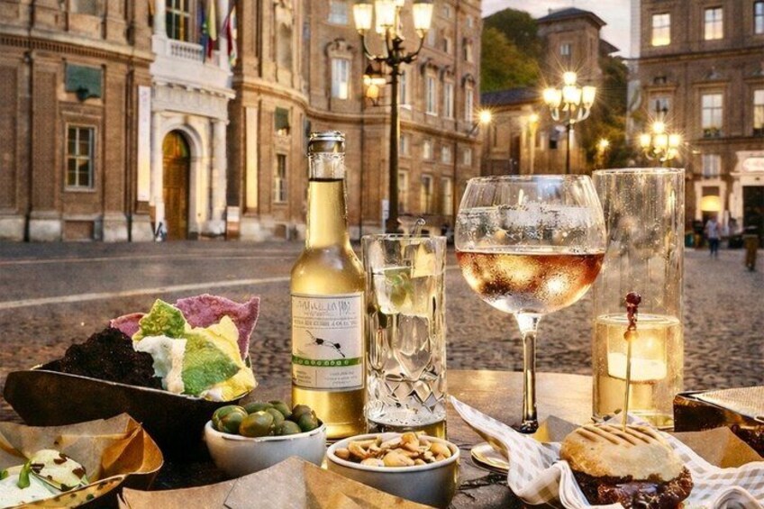 A table with food and drinks, a bottle of wine, and a glass of red wine in front of a historic building. 1