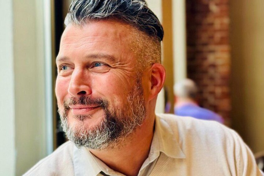 A person with a beard and short hair, wearing a light-colored shirt, sitting indoors. 4