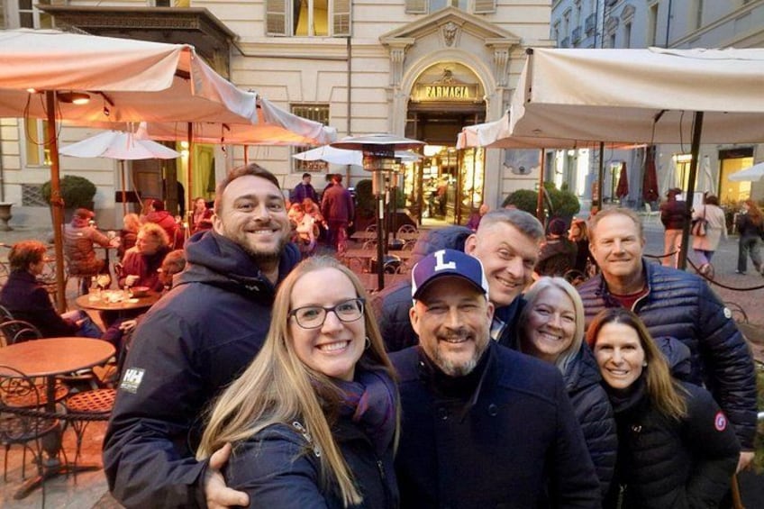 A group of people gathered around outdoor tables with umbrellas, in front of a building with a sign reading 'PARMAGGIA.' 3