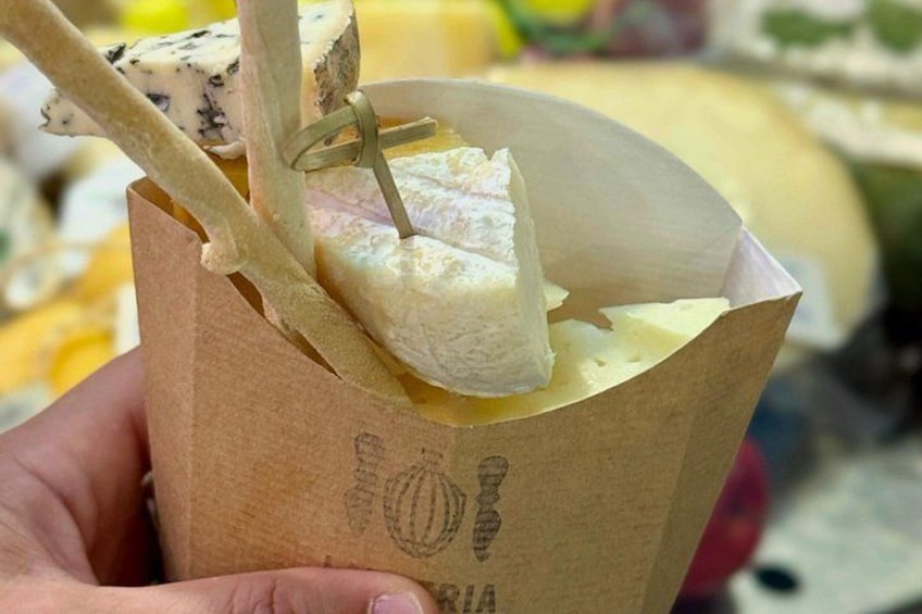 A hand holding a paper cup with a piece of cheese and a wooden skewer. 3