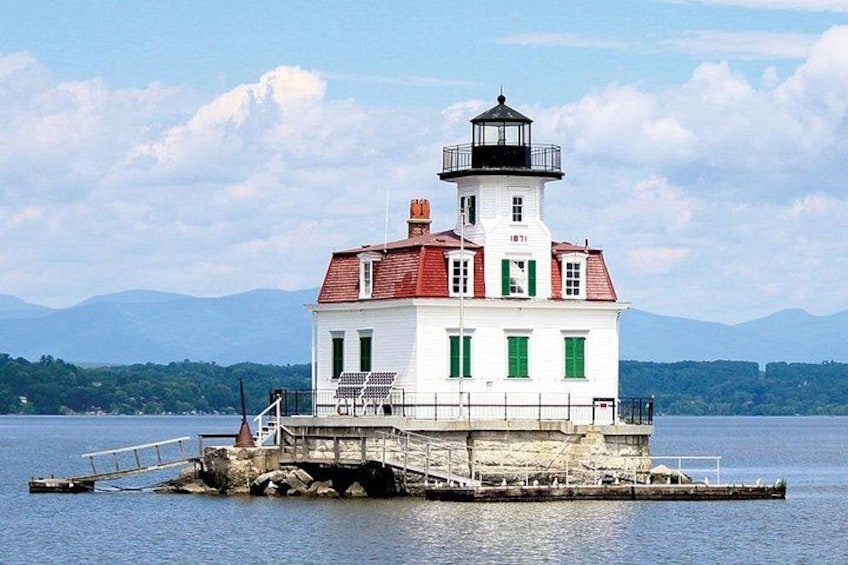 Un phare blanc, surmonté d’un toit rouge, se dresse sur une petite île, entouré d’eaux calmes et dominé par des montagnes lointaines. 2