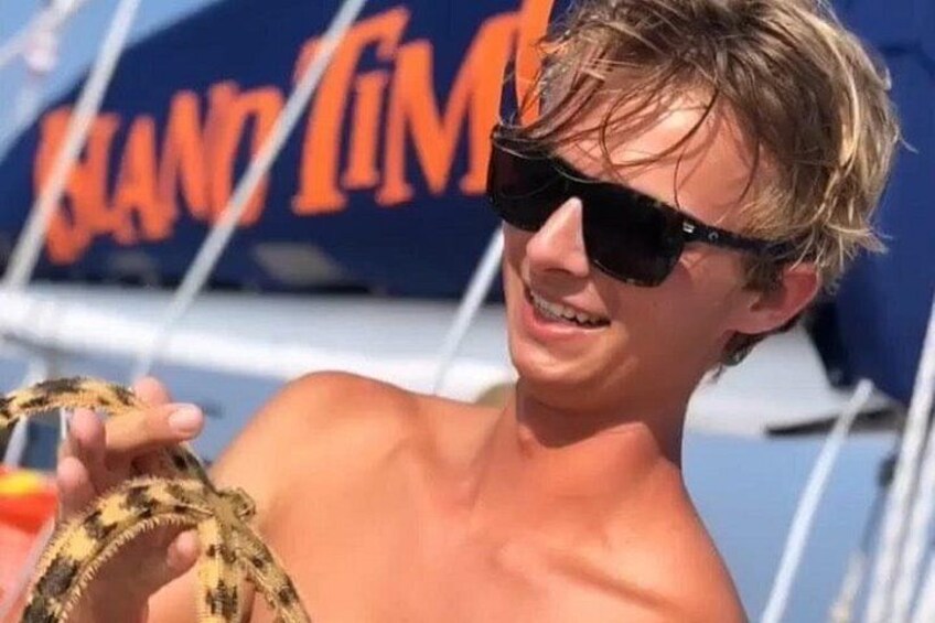 Una persona tiene un serpente su una barca, con un cartello 'Grand Tim' sullo sfondo. 2