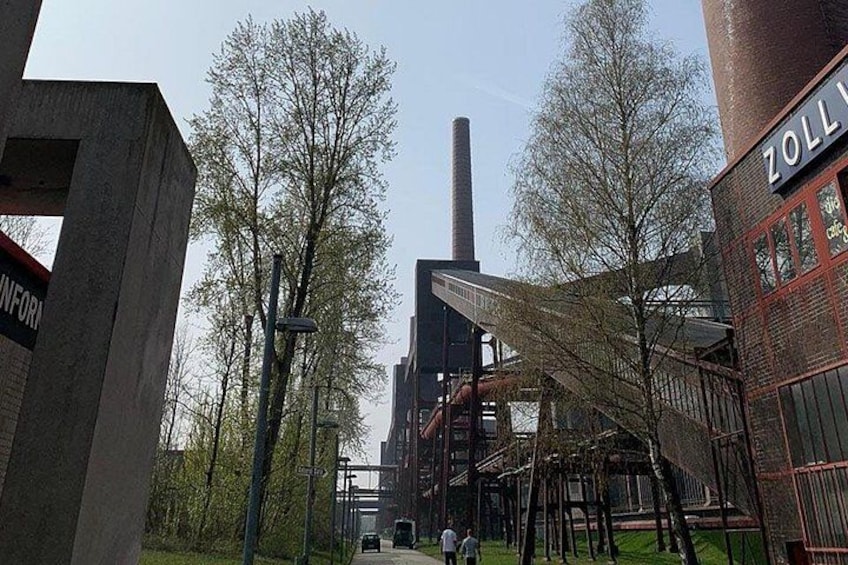 A large industrial building with a tall chimney, surrounded by trees and a pedestrian walkway. 4