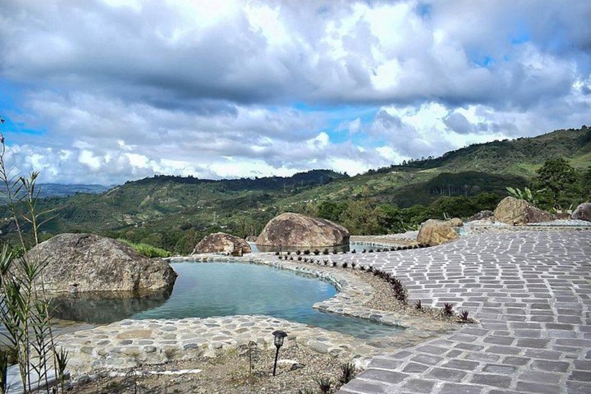 Ein steiniger Weg führt zu einem runden Pool, der von großen Steinen umgeben ist, mit einer hügeligen Landschaft im Hintergrund. 4