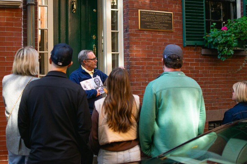 A group of people standing outside a brick building with a green door and a plaque. 2