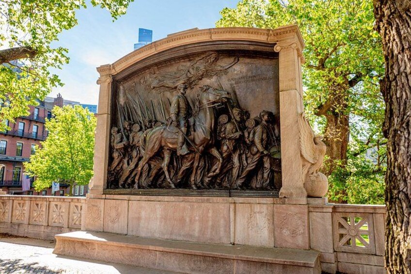 A bronze war memorial with relief sculptures, set in a park with trees and buildings in the background. 1
